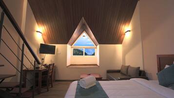 Un dormitorio con techo inclinado, una cama, un escritorio con silla, una mesita y una ventana con vista a una colina.
