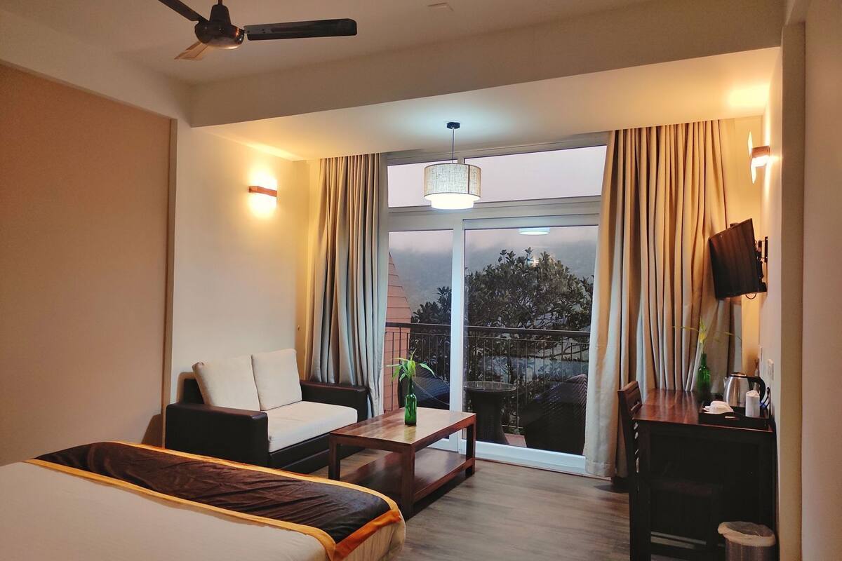 Ein Hotelzimmer mit Bett, Sofa, Couchtisch, Fernseher und einem Balkon mit Vorhängen.