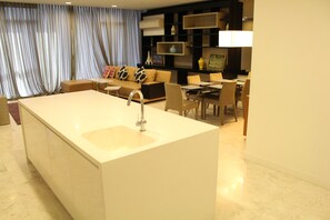 Een moderne woonkamer met een bank, eettafel en stoelen. In het midden staat een groot keukenblok met een spoelbak.