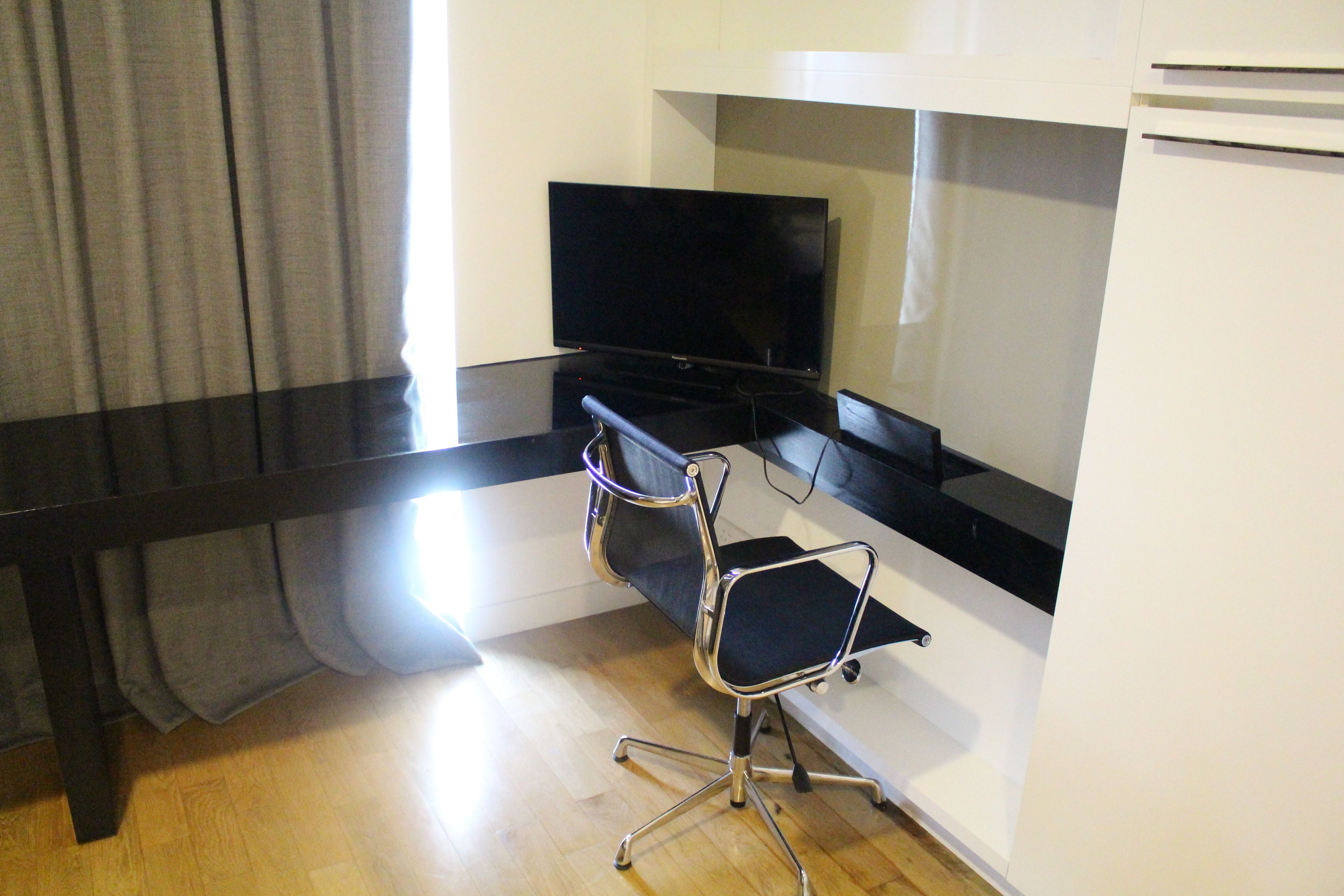 Una habitación con un escritorio negro, una silla negra, un televisor de pantalla plana y una pared blanca.