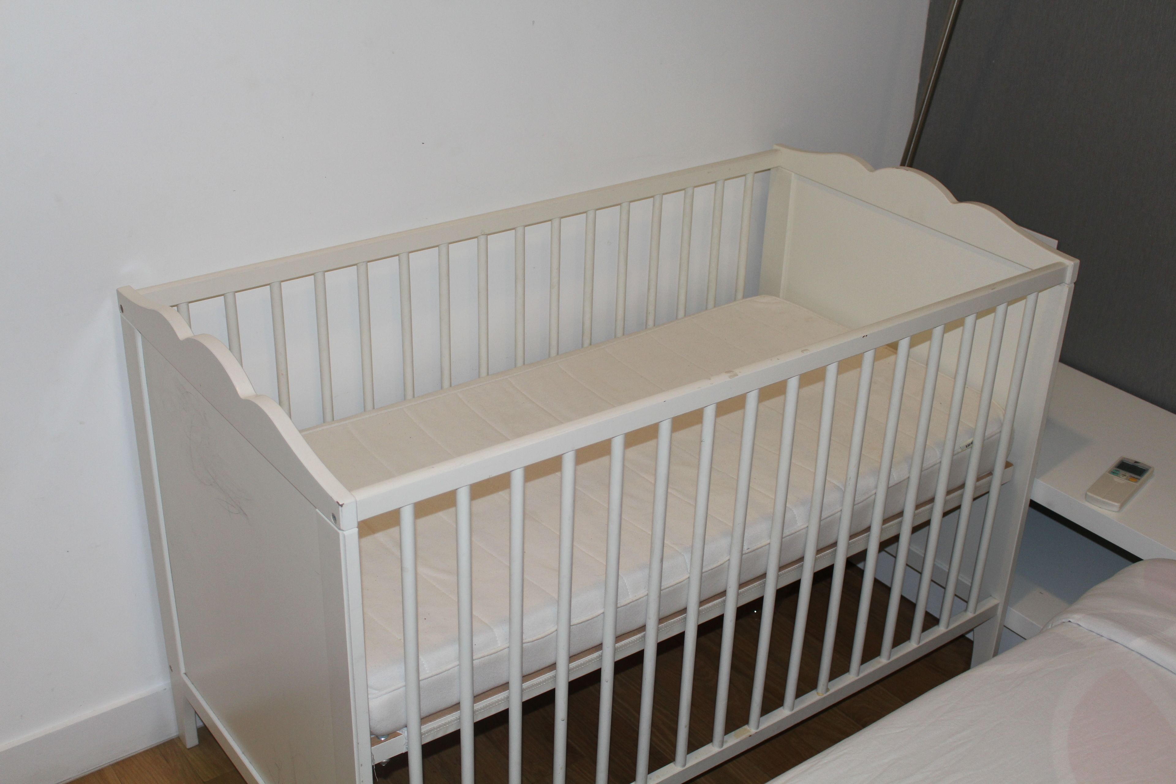 Ein weißes Kinderbett mit Matratze und Schlaufengittern, vor einer schlichten Wand platziert.