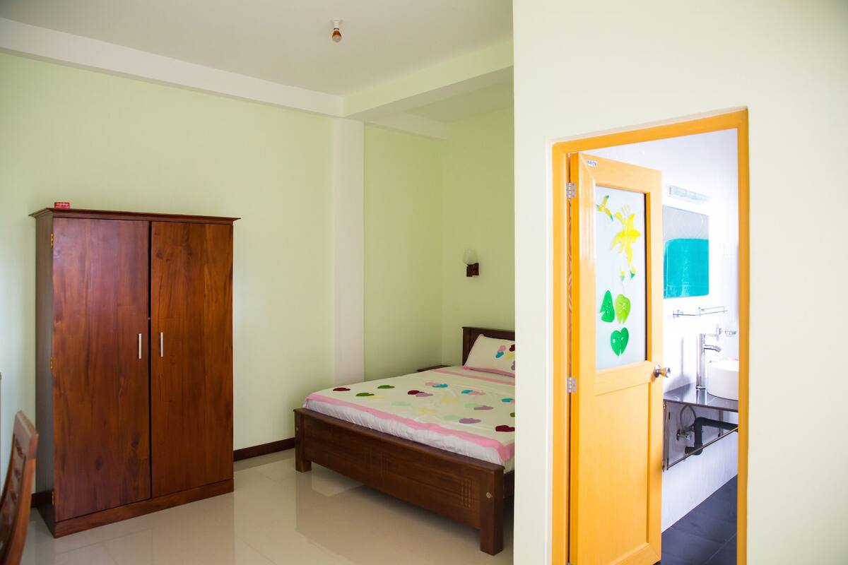 Un dormitorio con un armario de madera, una cama con una colcha colorida y un baño visible a través de una puerta abierta.