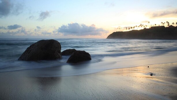 A beach with large rocks in the foreground and a distant view of palm trees and a sunset. - Horizon Inn (Weligama)