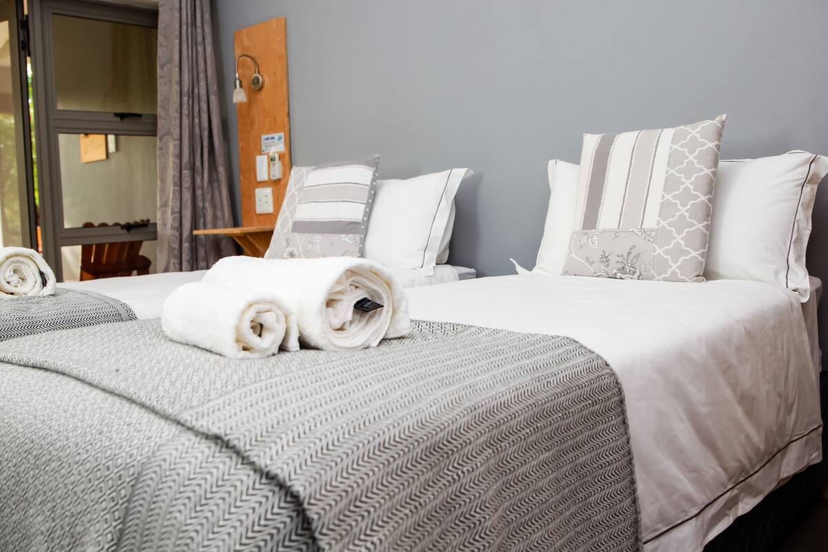 Una cama bien tendida con toallas enrolladas, una manta gris estampada y almohadas blancas con diferentes diseños.