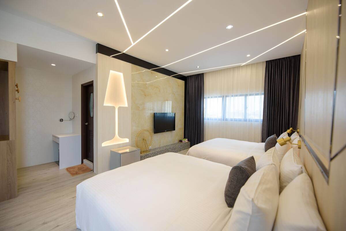 A modern hotel room with two beds, a TV mounted on the wall, and a small desk with a lamp.