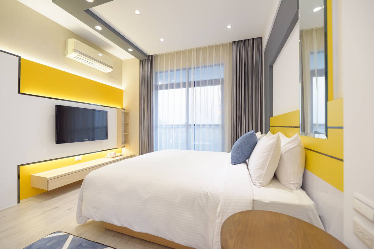 A modern hotel room with a large bed, a flat-screen TV mounted on the wall, and a large window with curtains.