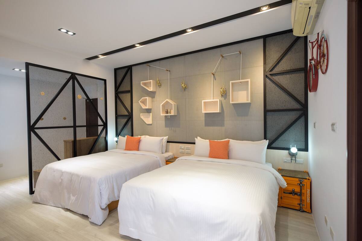 A modern hotel room with two beds, a large wall-mounted TV, and a small table with a lamp.