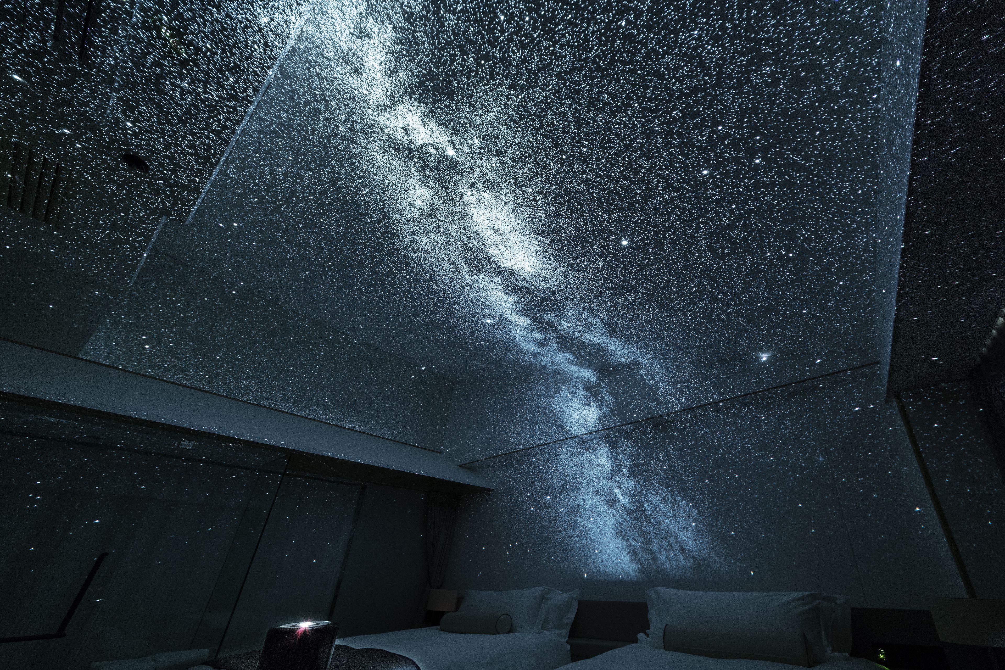 Een kamer met een bed, een nachtkastje en een projectie van een sterrenhemel op het plafond.