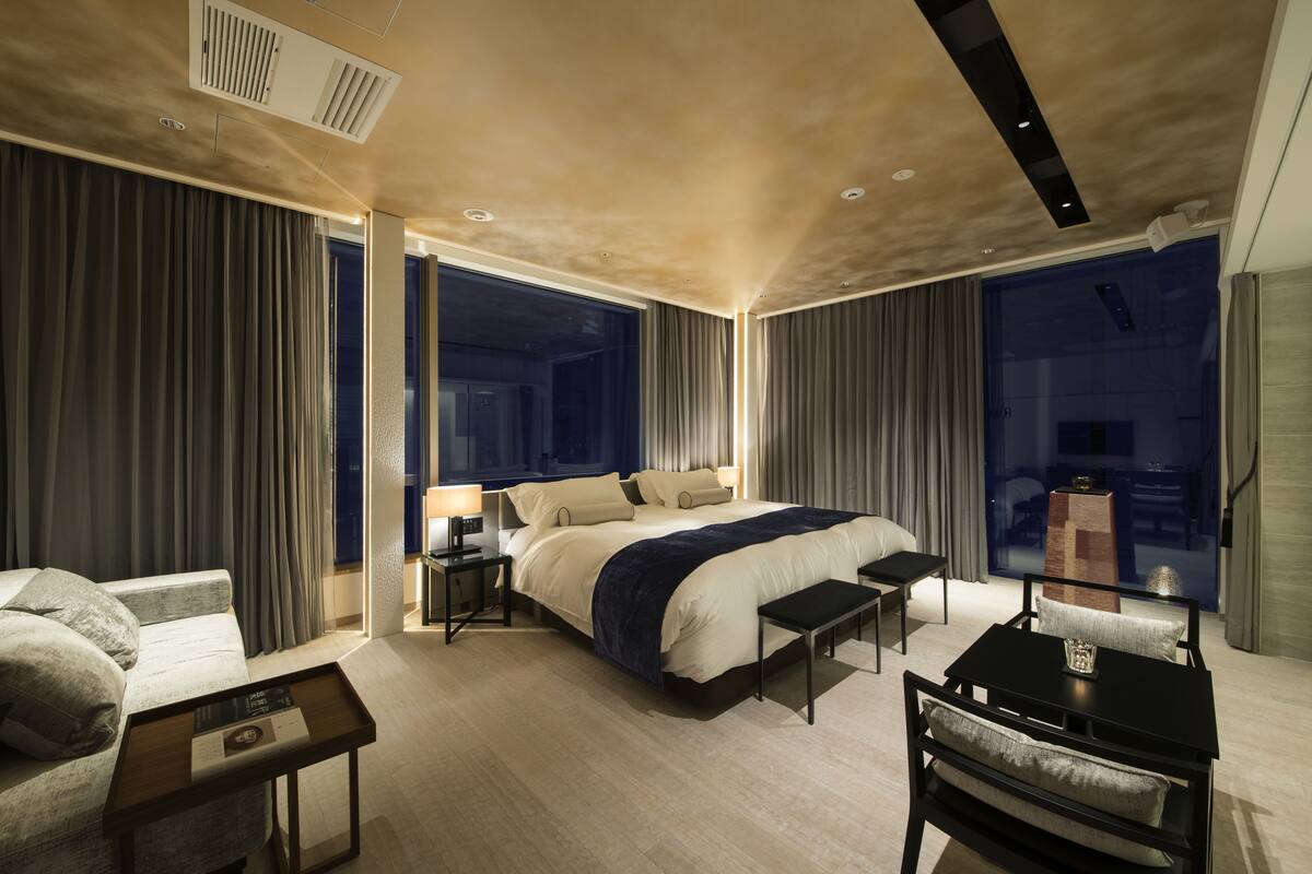 Ein modernes Hotelzimmer mit einem großen Bett, einem Schreibtisch und einem Stuhl. Es gibt ein Fenster mit Vorhängen, einen Nachttisch und eine Lampe.