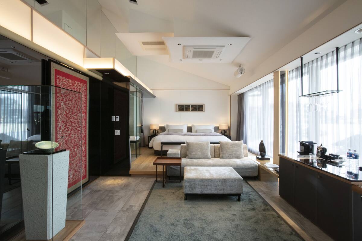 Ein modernes Hotelzimmer mit einem großen Bett, einer Sitzecke mit Sofa und Hocker, einer kleinen Küchenzeile und großen Fenstern mit Blick.