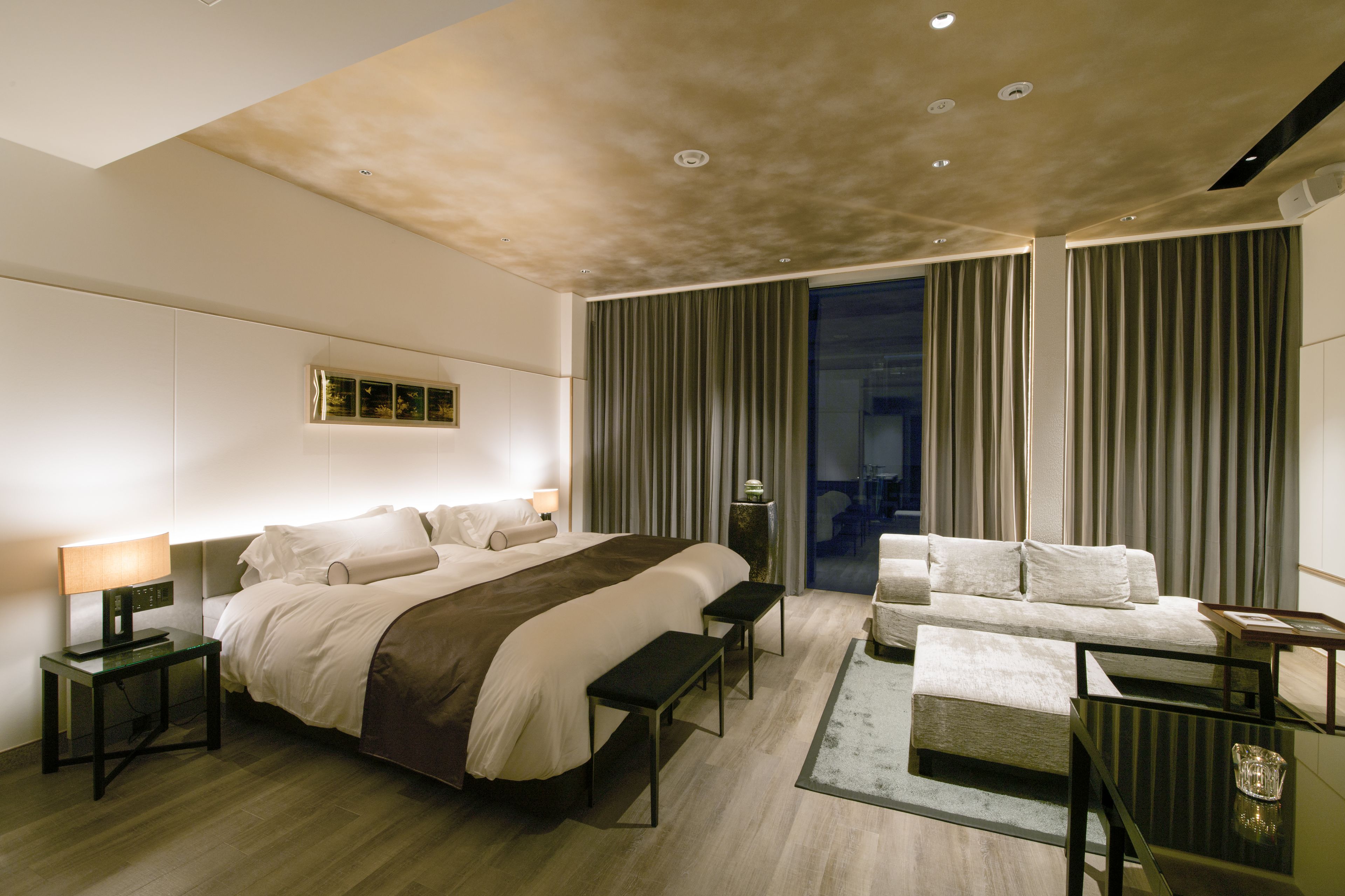 A modern hotel room with a large bed, a sofa, a desk, and a view of the city through the window.