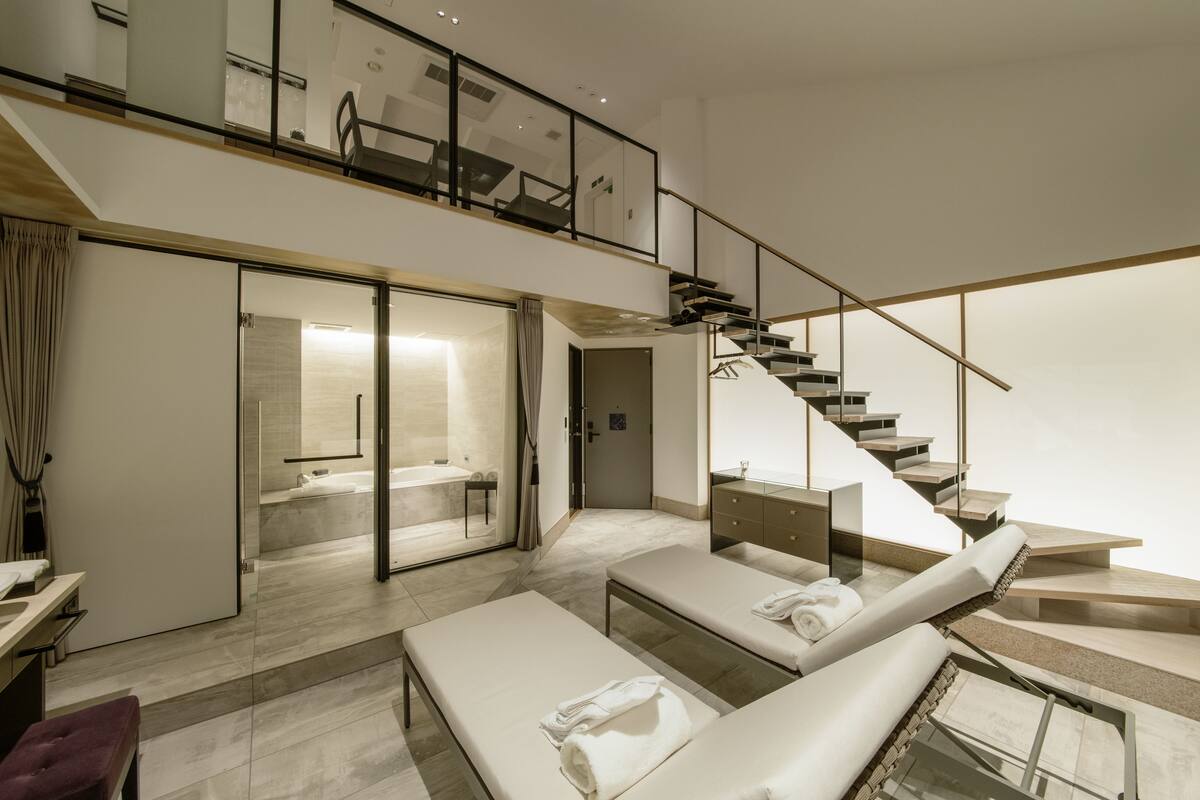 Ein modernes Hotelzimmer mit großzügigem Wohnbereich, einem großen Fenster und einer Treppe, die zu einer oberen Etage führt.