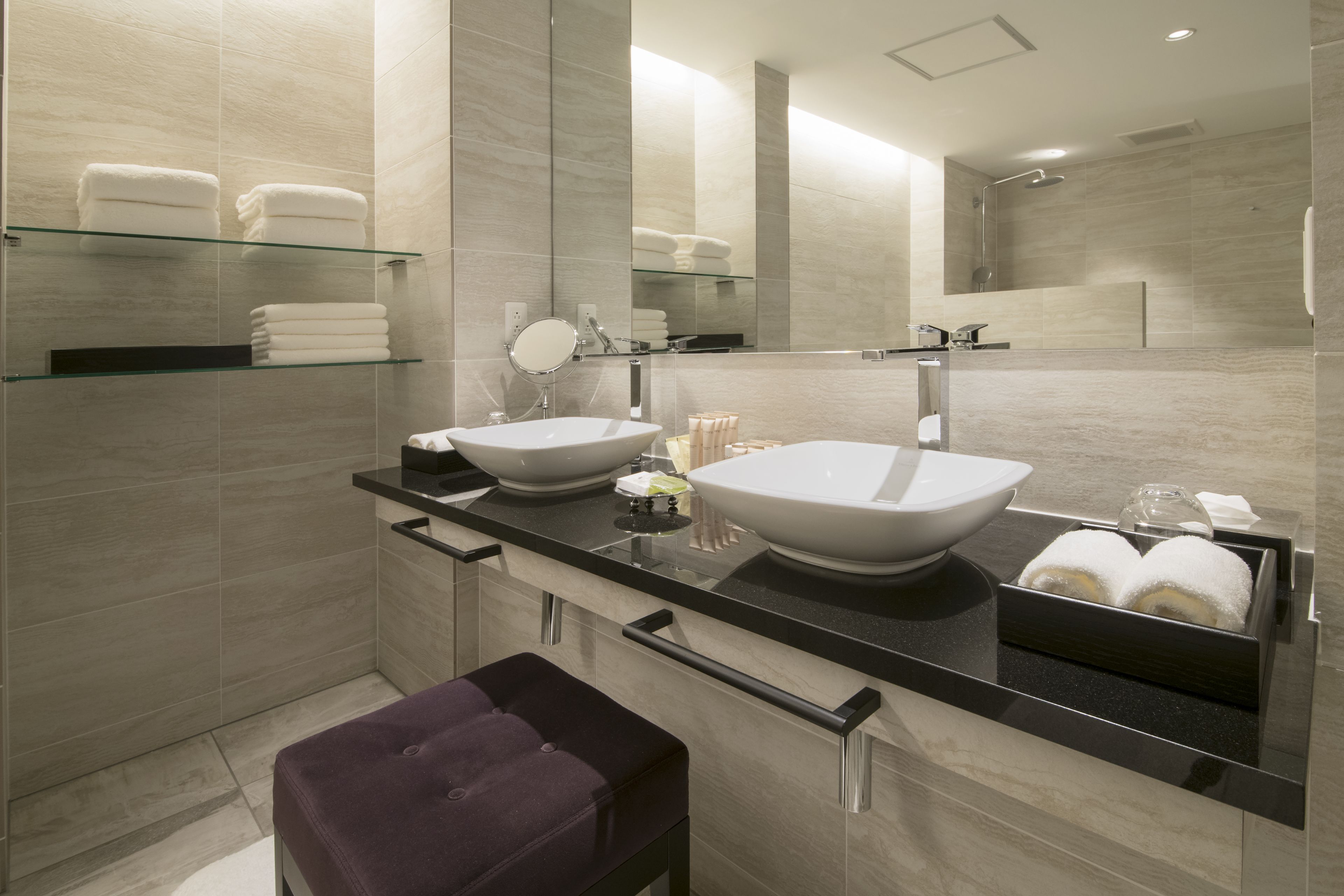 A modern bathroom with two large sinks, a glass shelf with towels, a vanity mirror, and a bench.