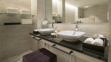 A modern bathroom with two large sinks, a glass shelf with towels, a vanity mirror, and a bench.