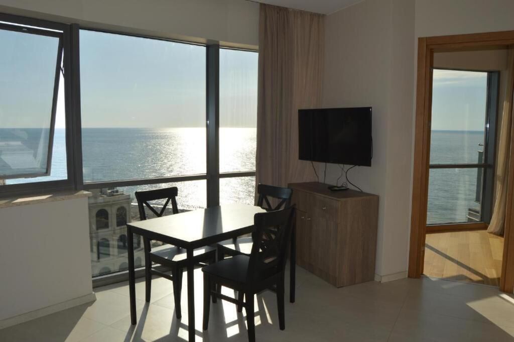 A room with a dining table, chairs, a TV, and large windows overlooking the sea.