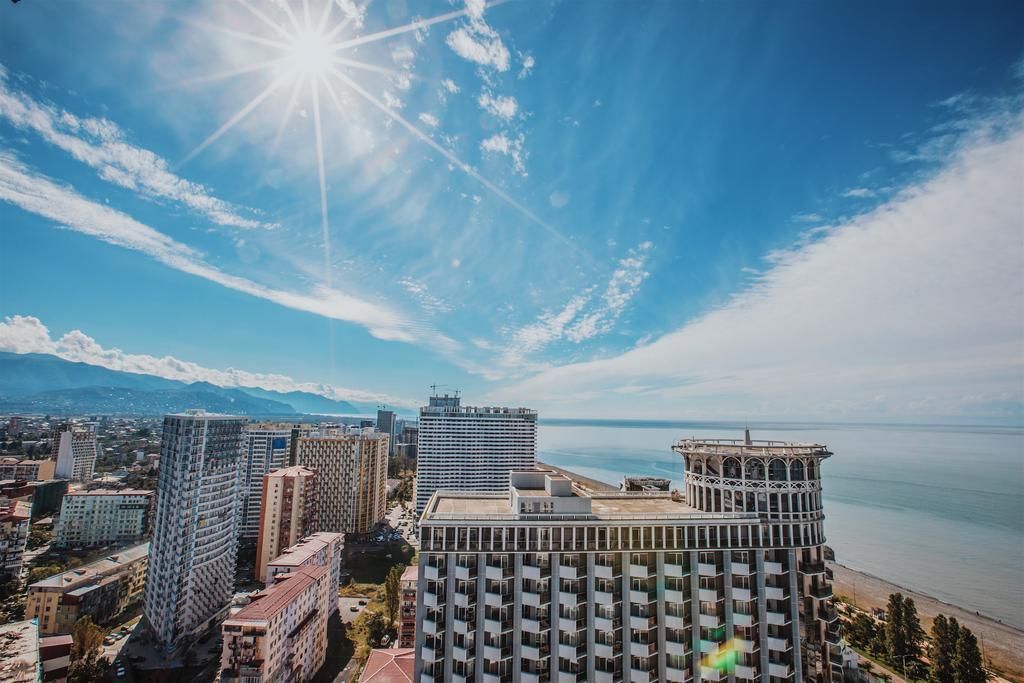 A high-rise building with a modern architectural design, located near a coastline with a clear sky and sun.