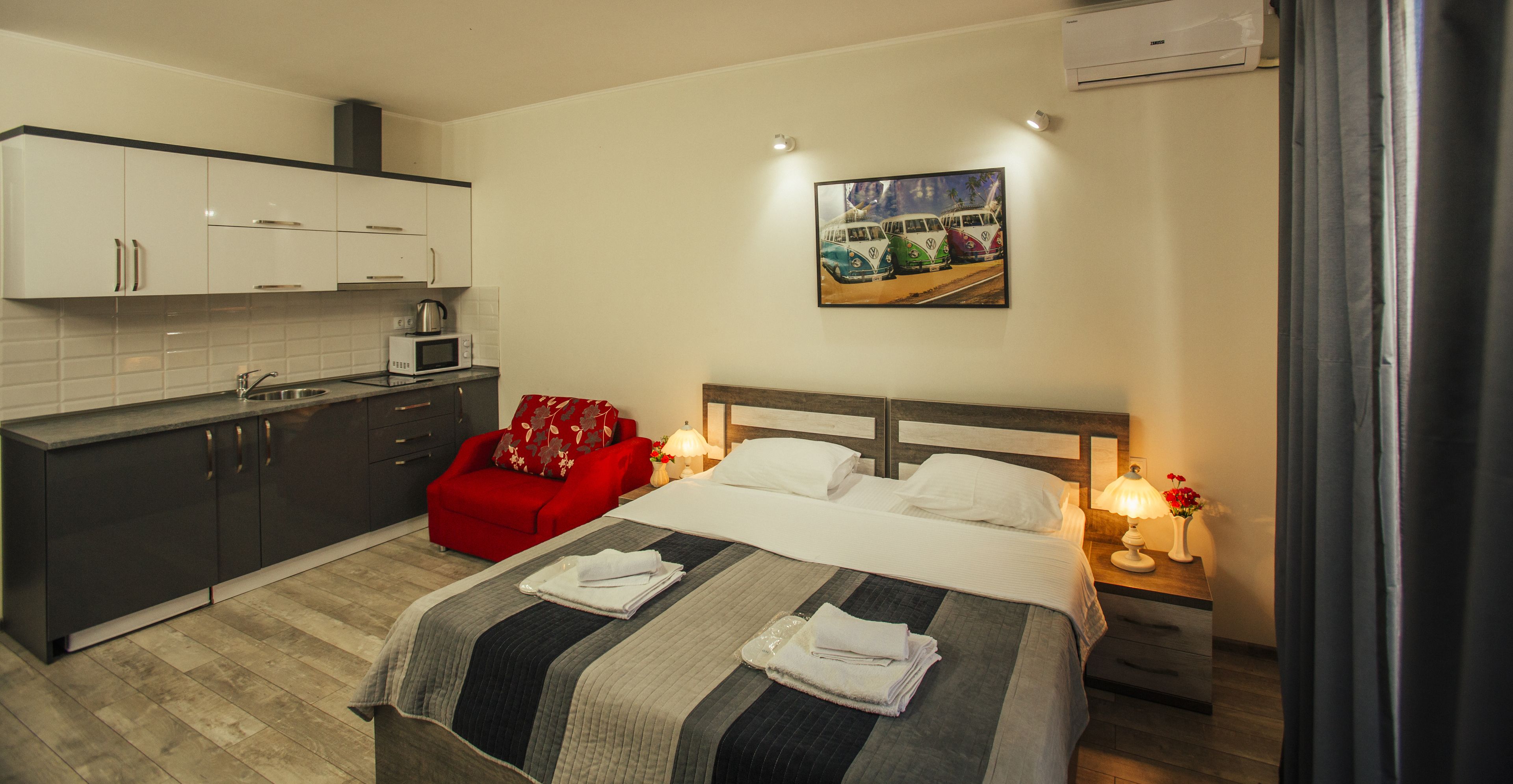 Una habitación de hotel con una cama, una silla roja, una zona de cocina y un cuadro en la pared.