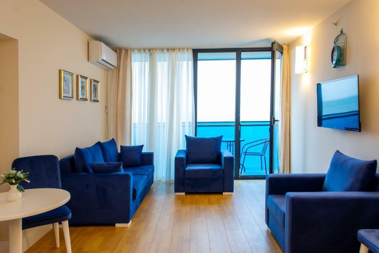 A modern living room with a sofa set, a coffee table, and a TV. There is a balcony with a view of the sea.