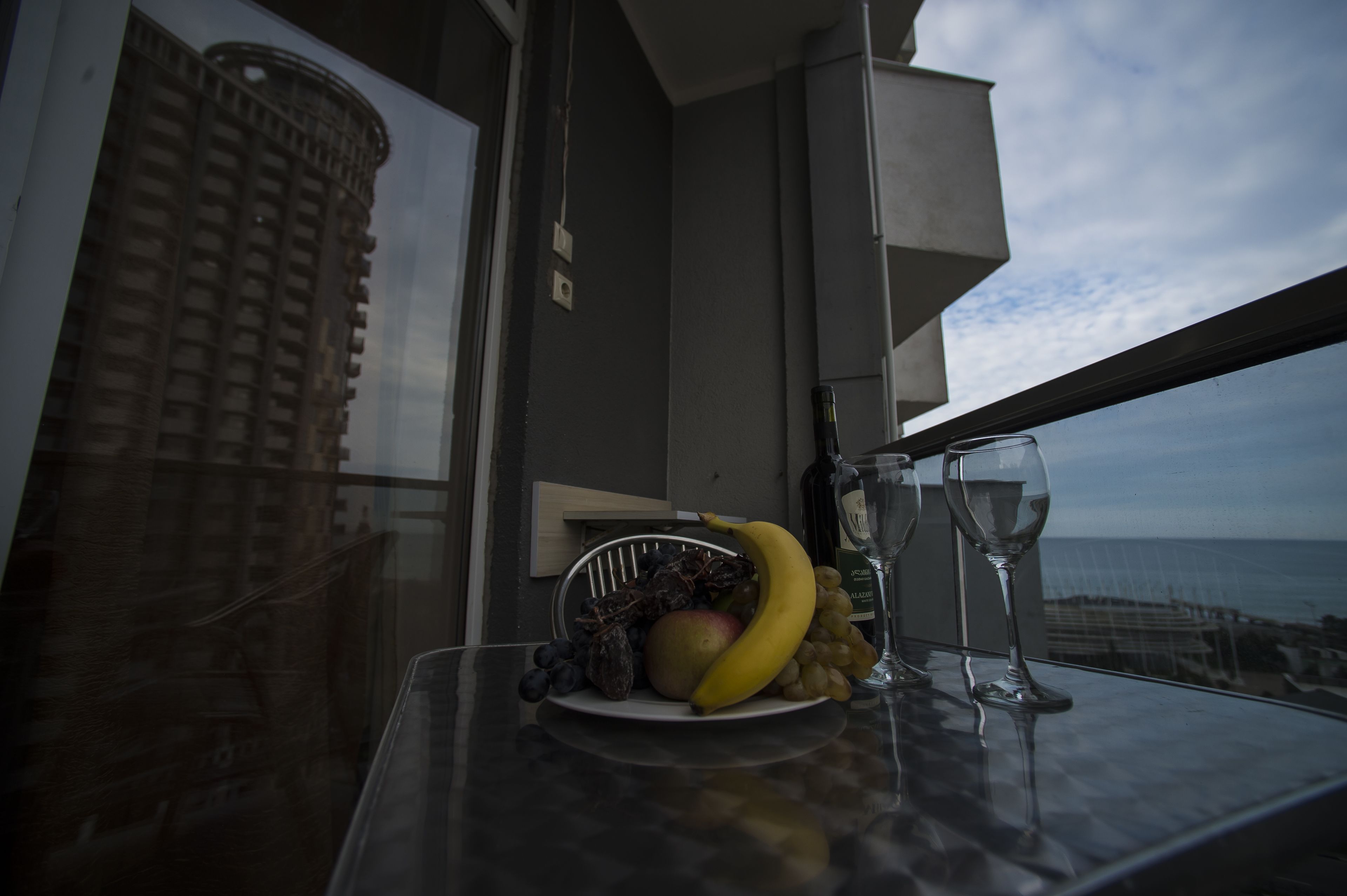 A table with a fruit basket, a bottle, and two wine glasses on a balcony overlooking the sea.