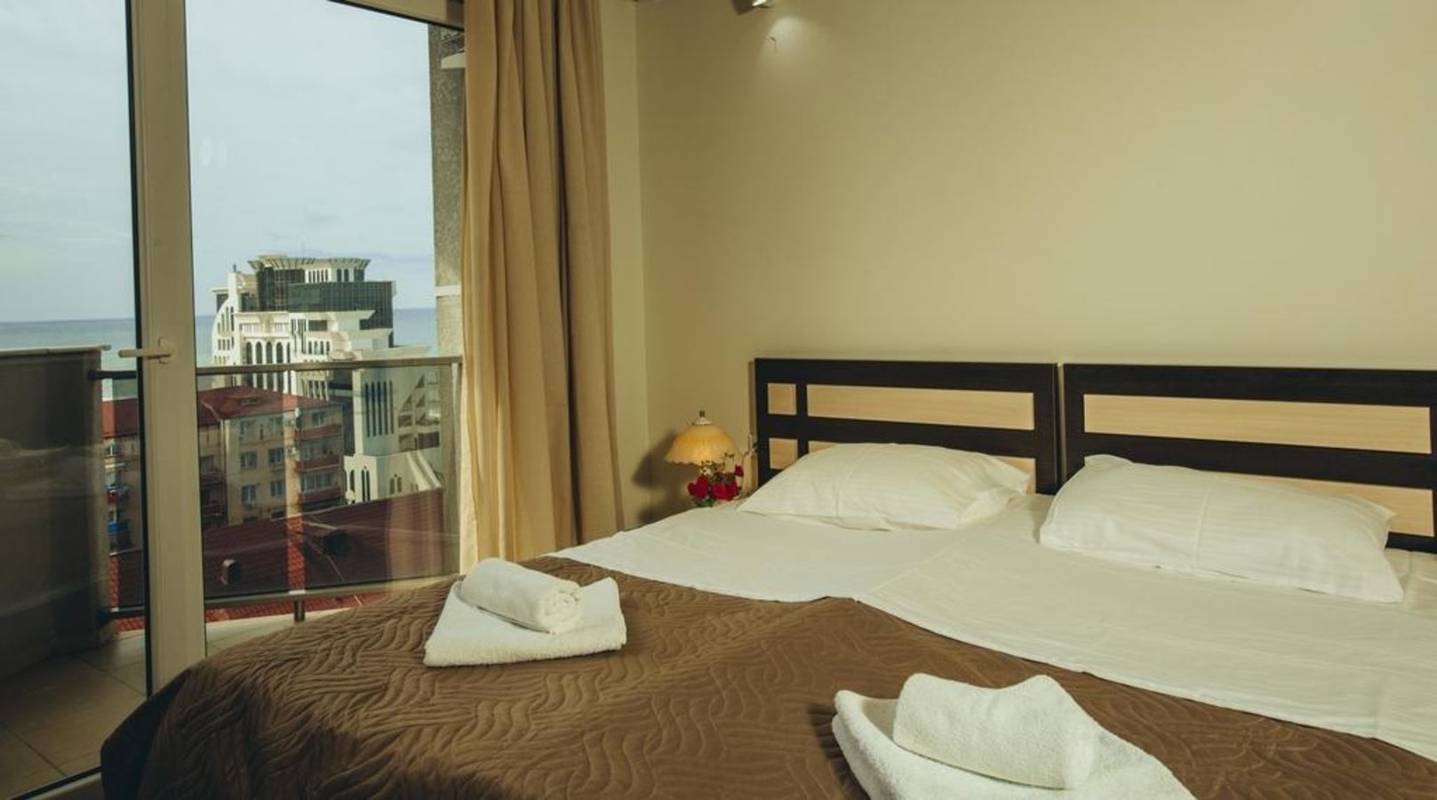Habitación de hotel con una cama grande, dos almohadas, una lámpara de noche y vistas a edificios a través de una puerta de cristal.