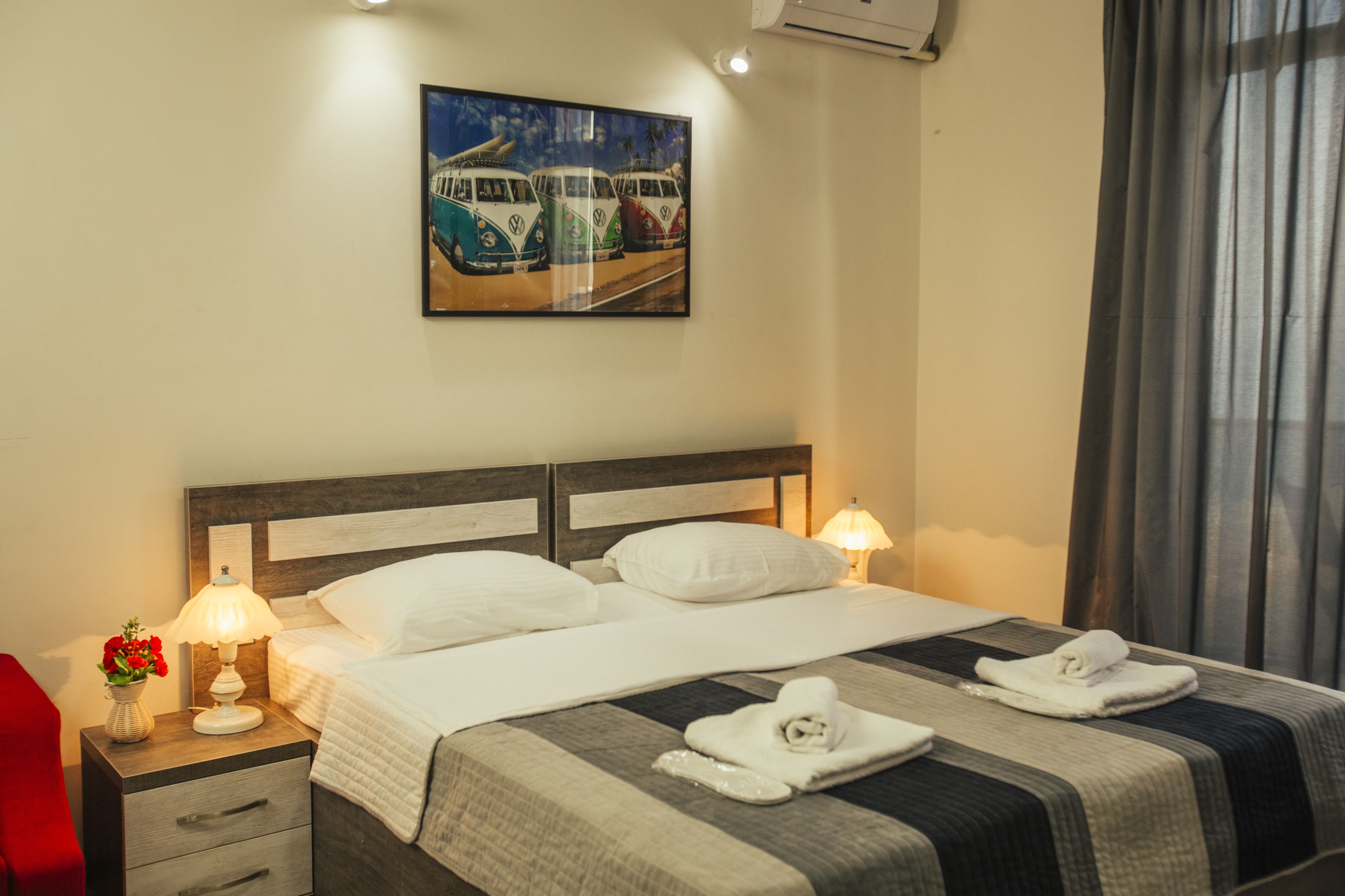 A hotel room with a bed, two bedside lamps, a nightstand with a vase of flowers, a painting of vintage cars on the wall, and a window with curtains.