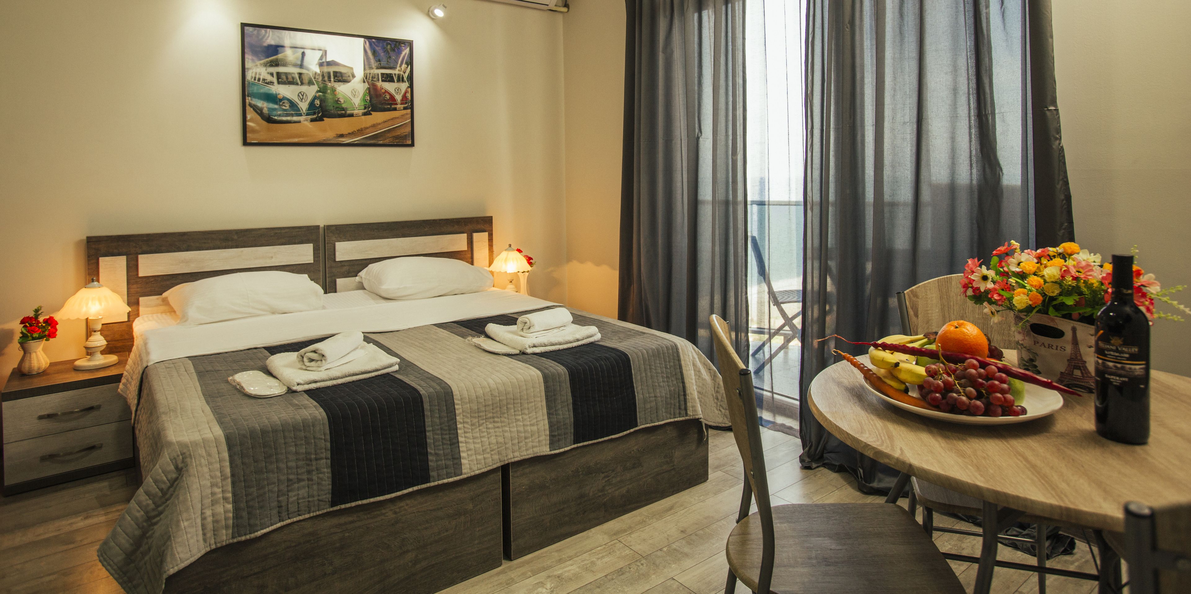 A hotel room with a bed, bedside tables, a dining table with fruit and a bottle, and a view through the window.