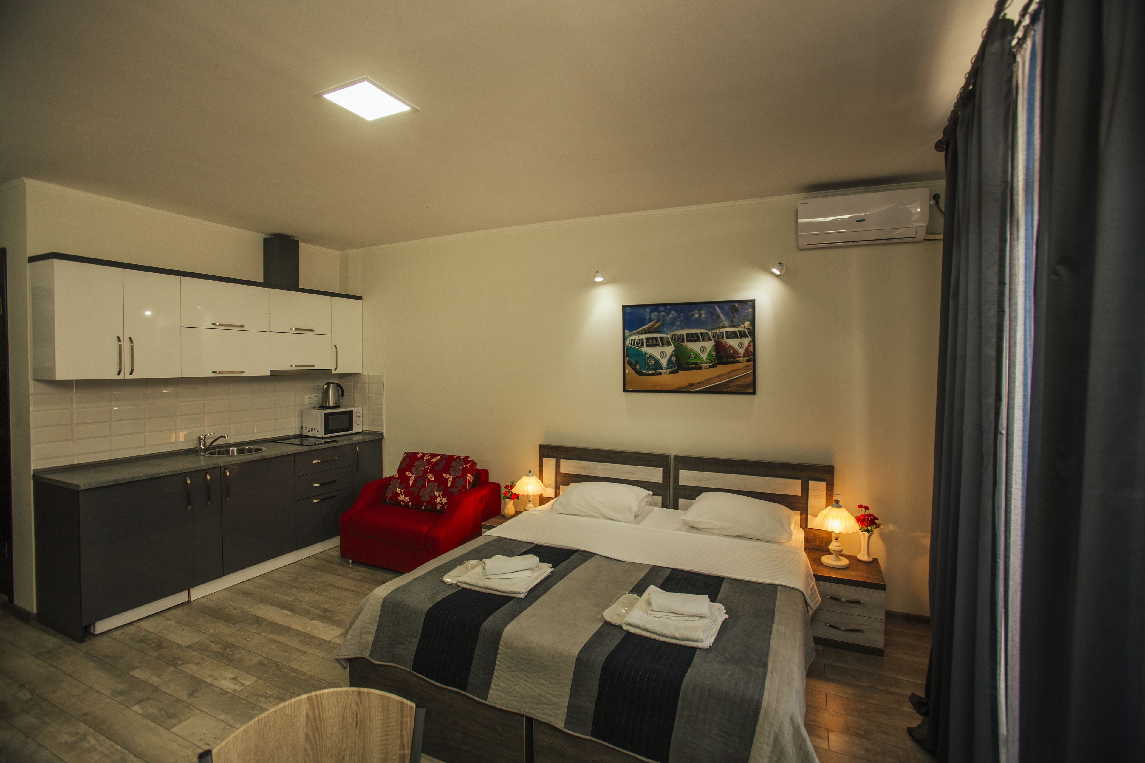 Una habitación de hotel moderna con kitchenette, una cama y toallas ordenadas, una silla roja y un cuadro en la pared.