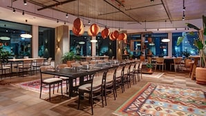 Un vaste espace dédié à la salle à manger, avec de longues tables et des chaises, des luminaires suspendus et de grandes fenêtres offrant une vue sur le paysage urbain.