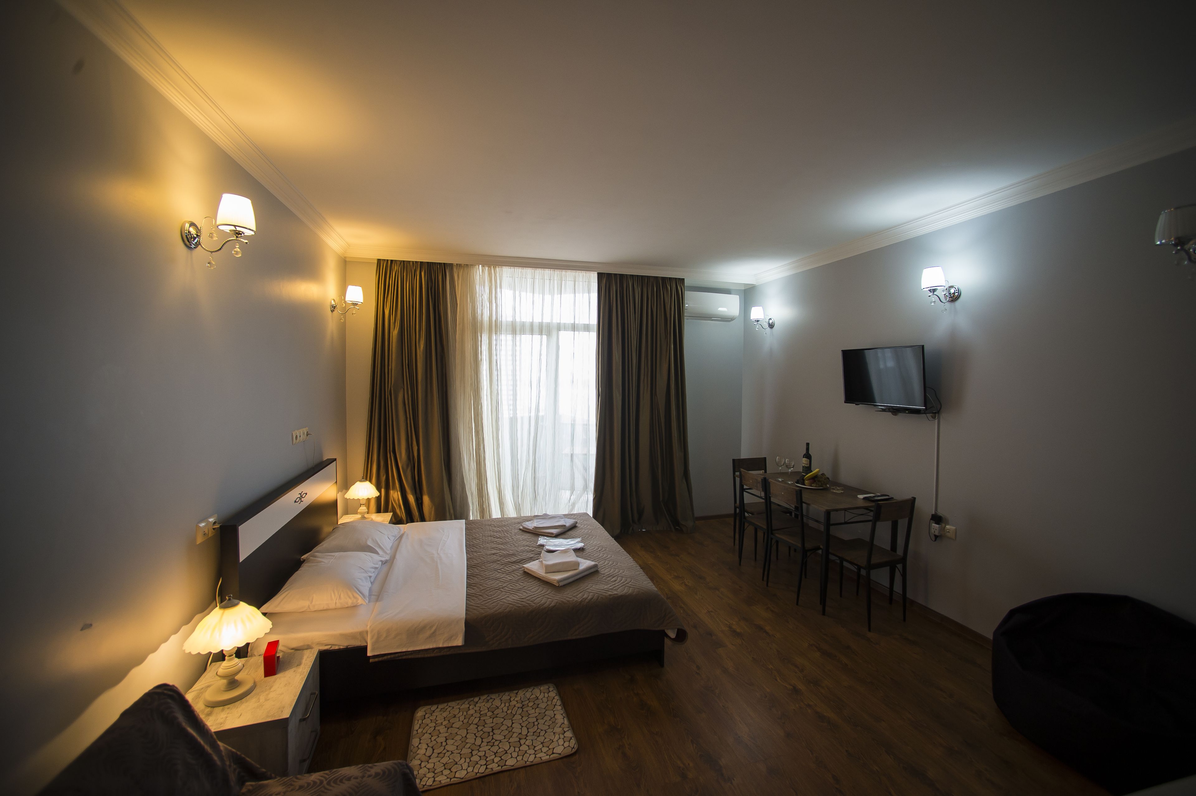 A hotel room with a bed, bedside table, lamp, curtains, a dining table with chairs, and a TV on the wall.