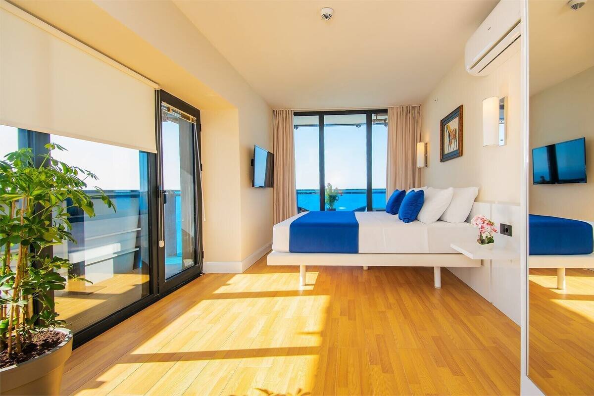 Ein modernes Hotelzimmer mit Bett, Fernseher, Balkon mit Pflanze und Blick auf das Meer.