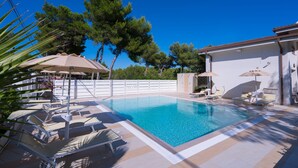 A swimming pool with lounge chairs and umbrellas, surrounded by trees and a white building.