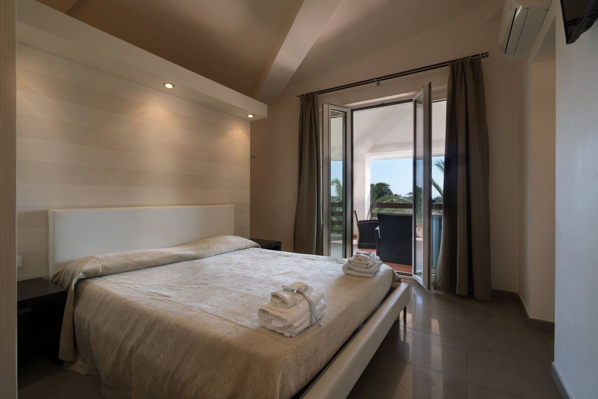 A modern hotel room with a large bed, a nightstand, a chair, and a view of the outdoors through a sliding glass door.
