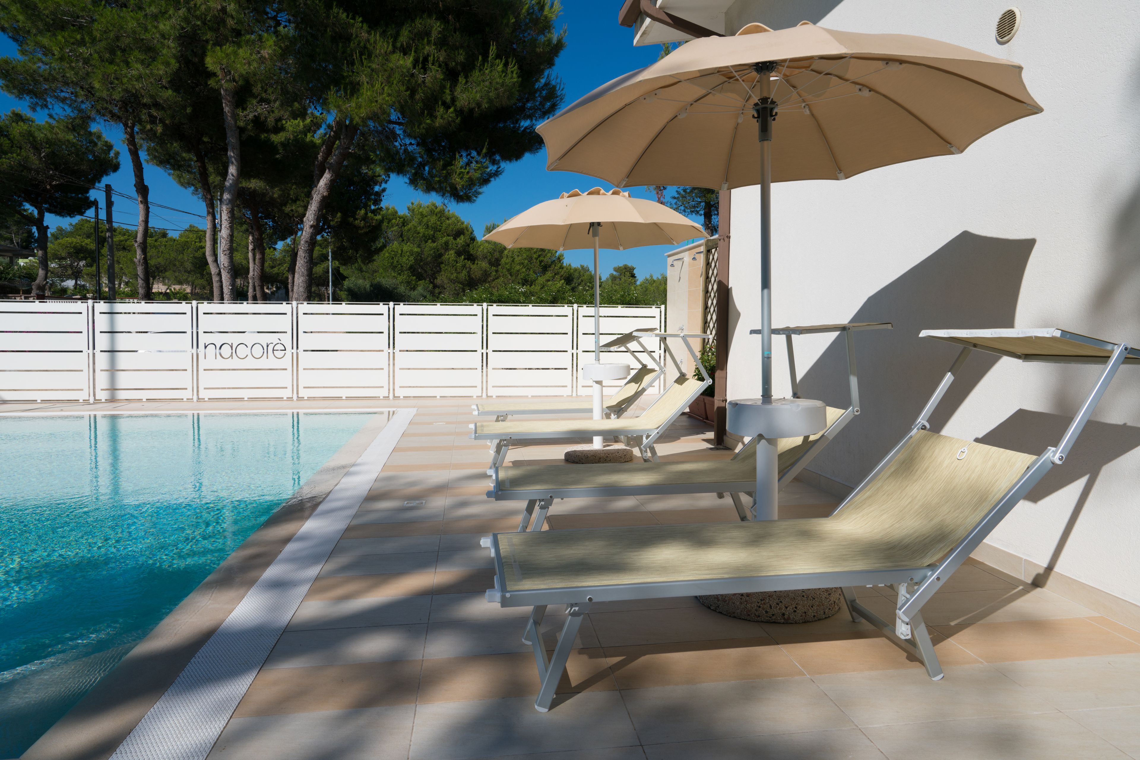 Área à beira da piscina com espreguiçadeiras e guarda-sóis, um deck revestido com azulejos e um cercado branco com árvores ao fundo.