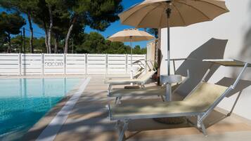 Área à beira da piscina com espreguiçadeiras e guarda-sóis, um deck revestido com azulejos e um cercado branco com árvores ao fundo.