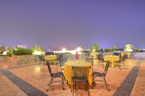 Outdoor dining area with round tables, black chairs, and potted plants on terracotta tile flooring.