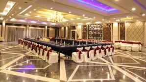 A banquet hall with a long table set for a formal event, surrounded by chairs with white and red sashes. - Hotel Chanakya (Patna)