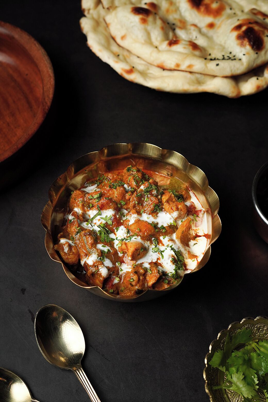 A bowl of chicken curry garnished with herbs and cream, accompanied by naan bread and a spoon on a dark surface.