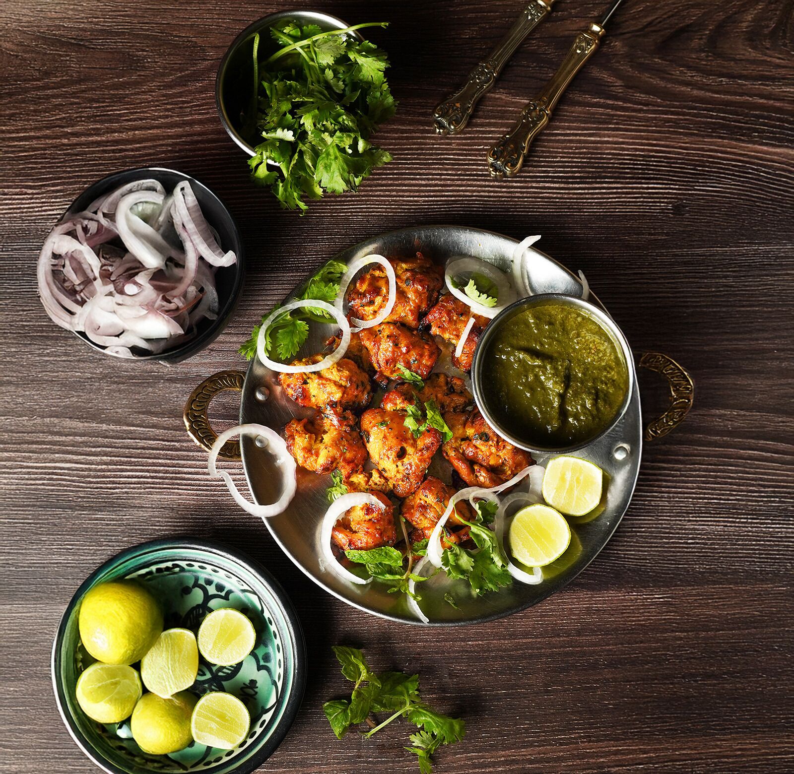 A plate of tandoori chicken with lemon wedges, onions, and a green chutney on a wooden table.