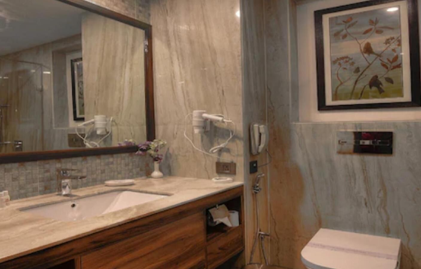 Un baño con lavamanos de mármol, tocador de madera, un espejo grande, un cuadro floral enmarco y un perchero para toallas.