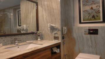 Un baño con lavamanos de mármol, tocador de madera, un espejo grande, un cuadro floral enmarco y un perchero para toallas.