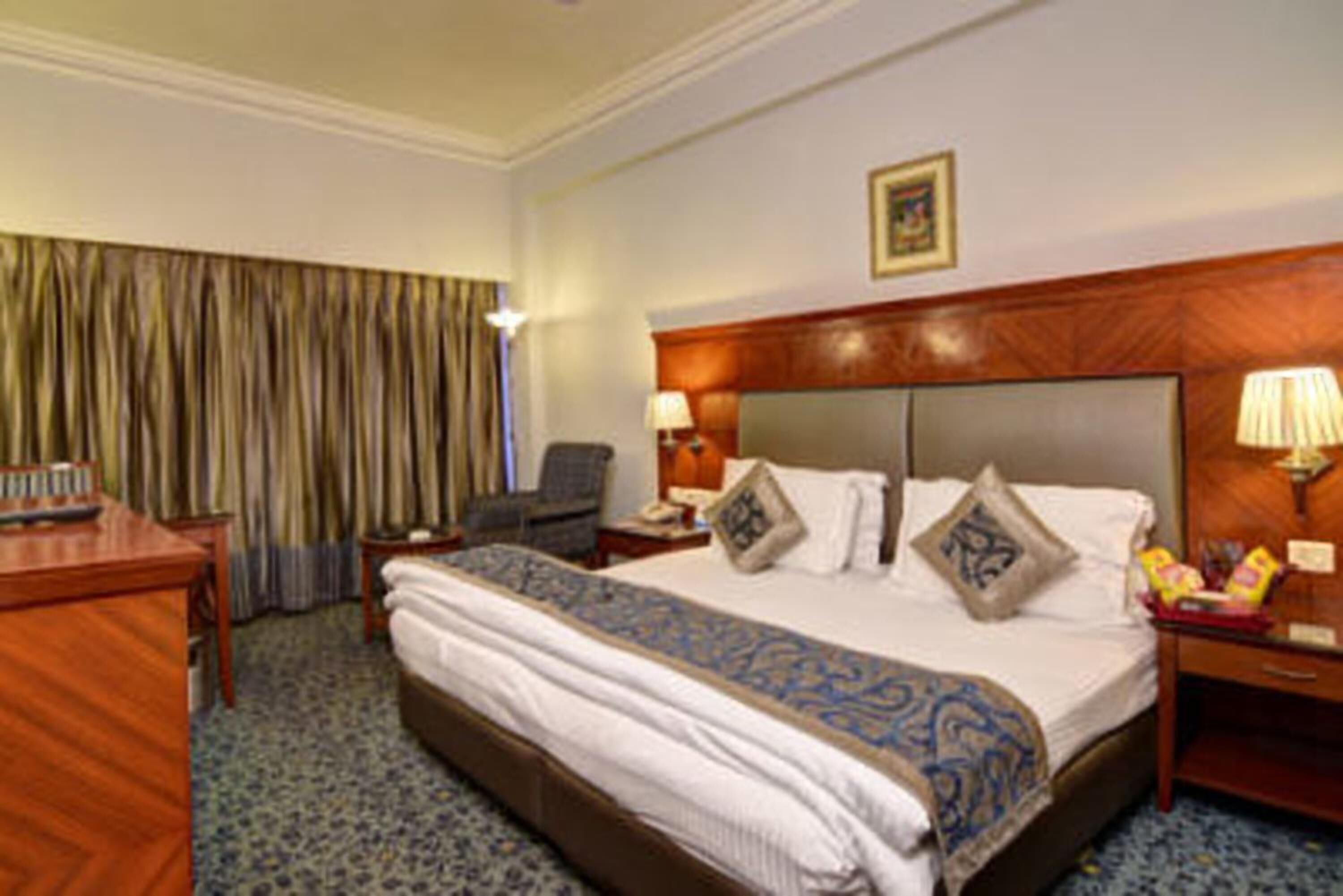 A hotel room with a large bed, a desk, a chair, a nightstand, and a lamp.