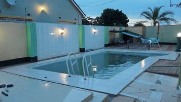 An outdoor swimming pool with a tiled deck, a ladder, and a covered seating area with chairs.
