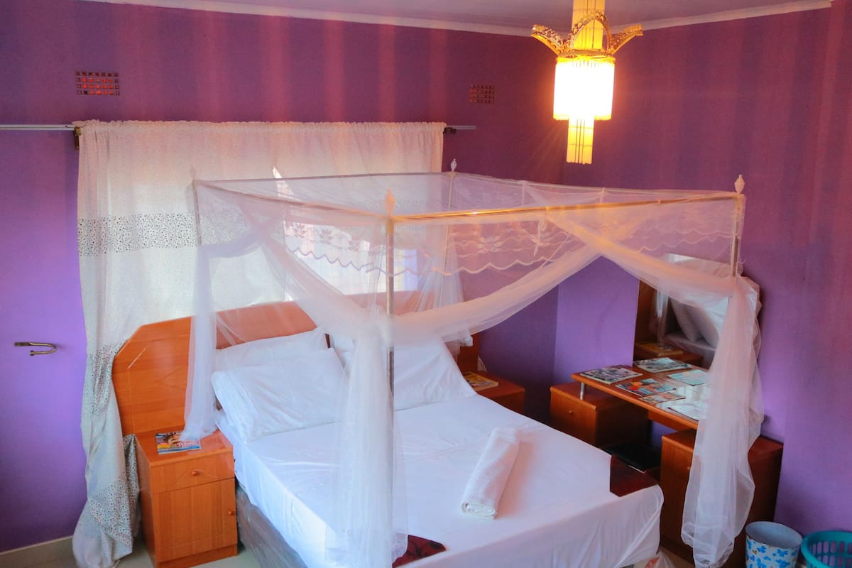 A single bed with a canopy, a bedside table, a nightstand, a desk with a chair, a trash bin, and a wall-mounted shelf.