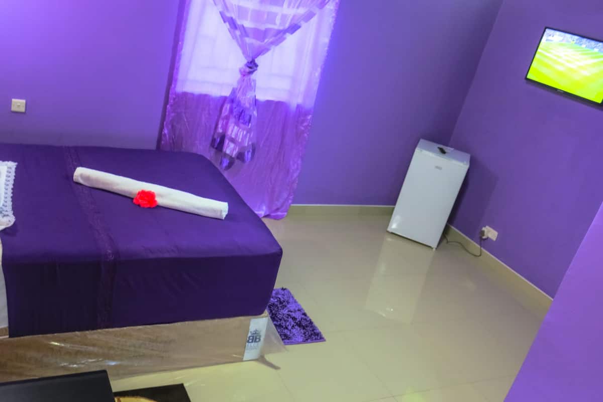 A room with a bed, a small table, a television on the wall, and a window with purple curtains.