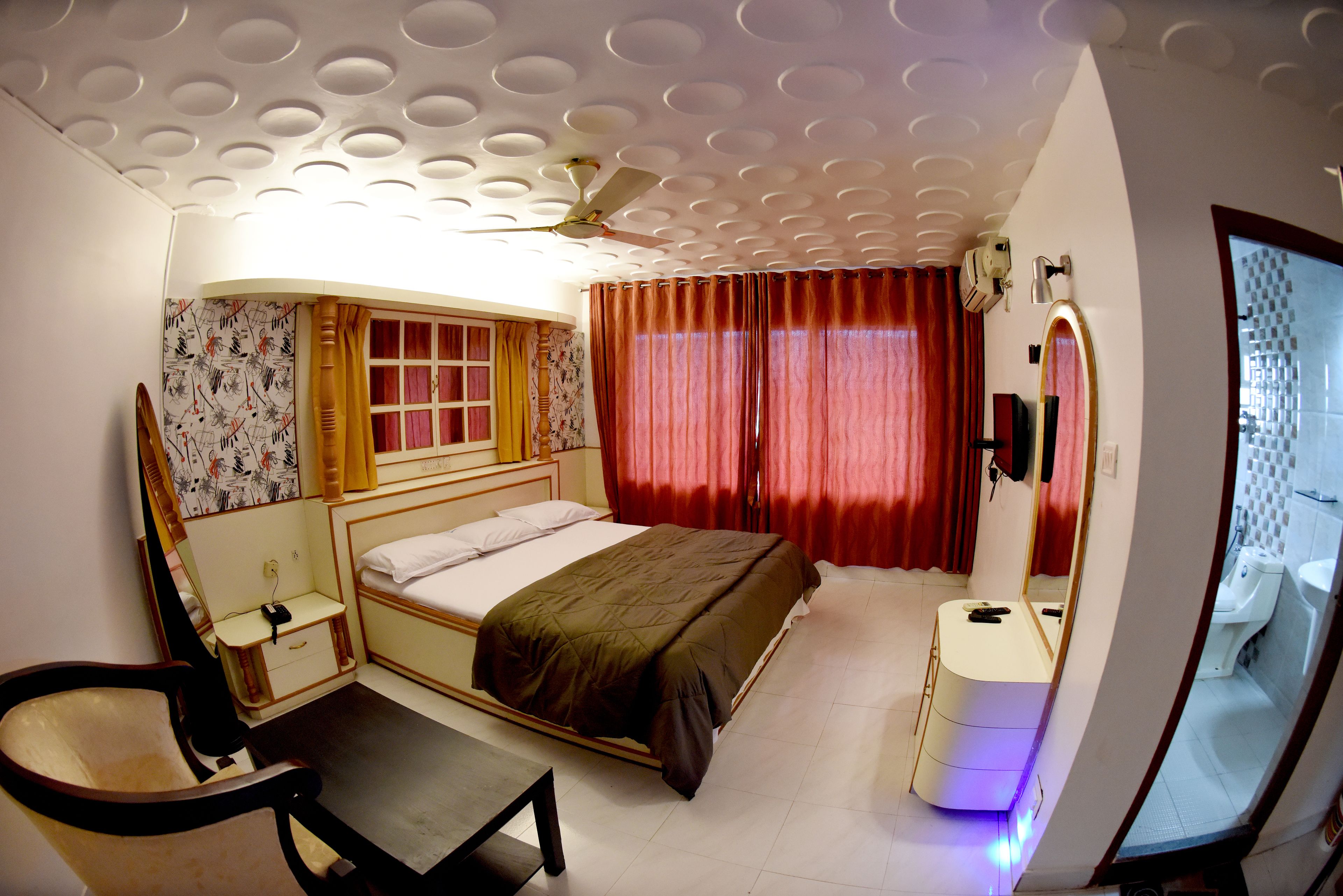Un dormitorio con cama, escritorio, silla, ventilador de techo, ventana con cortinas y baño con espejo y lavabo.