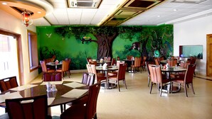 A restaurant with multiple tables and chairs, a green mural with trees and animals, and a ceiling with recessed lighting and air conditioning vents. - Sharada International Hotel (Udupi)