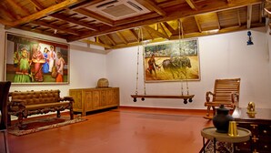 A spacious room with a high ceiling, wooden beams, a large painting, a swing, a bench, and a small table with a vase. - Sharada International Hotel (Udupi)