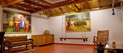 A spacious room with a high ceiling, wooden beams, a large painting, a swing, a bench, and a small table with a vase.