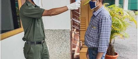 Una persona con uniforme verde realiza una toma de temperatura a otra persona que está afuera de un edificio con techo rojo y una maceta cerca.