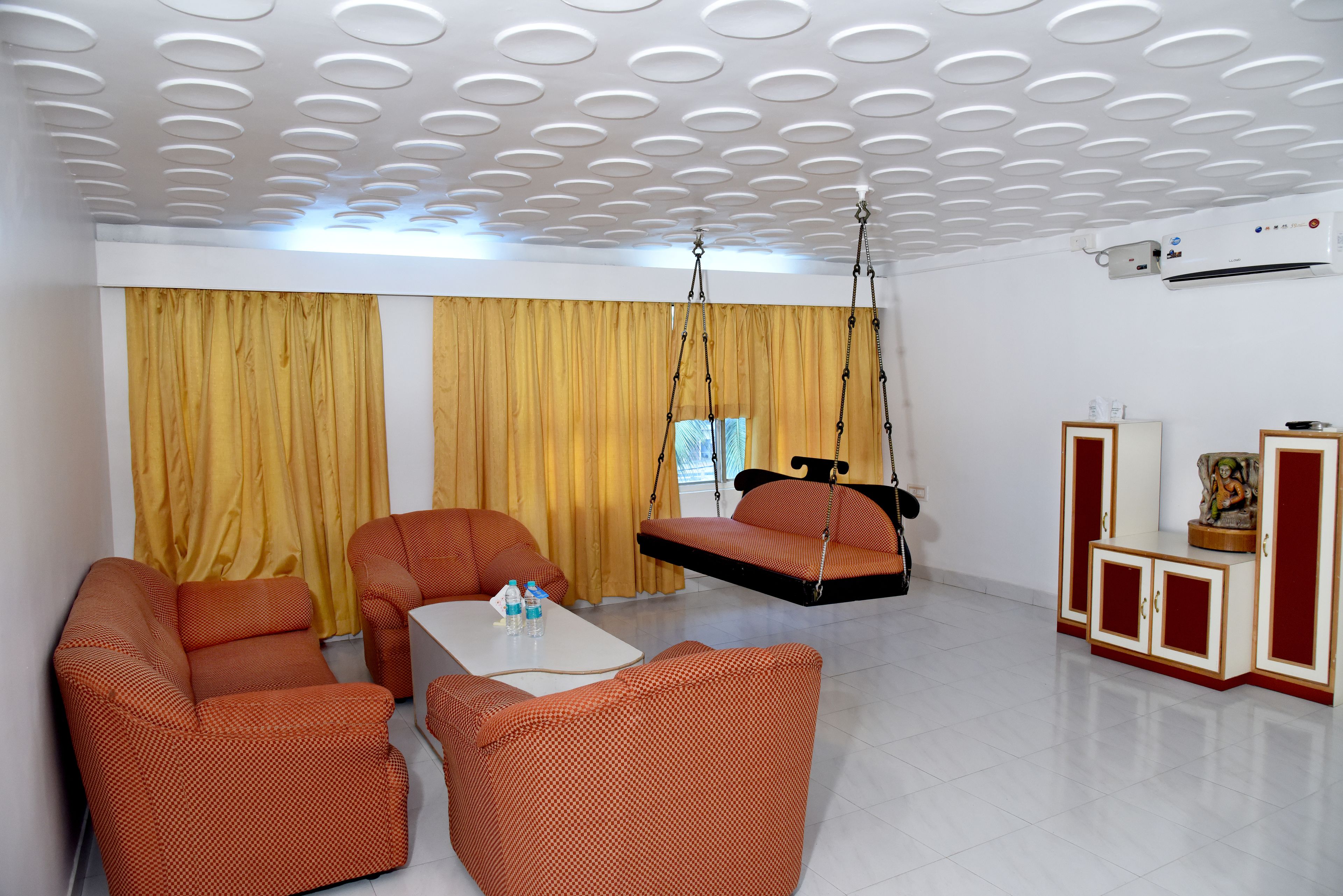 A modern living room with orange patterned sofas, a white coffee table, yellow curtains, and a ceiling with circular light fixtures.